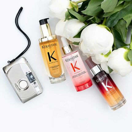 Drei Flaschen von Kerastase Haarpflegeprodukten (Elixir Ultime, Genesis und Nutritive) sind auf einer weißen Oberflache neben einer Vintage-Kamera und mehreren weißen Pfingstrosen mit grunen Blattern angeordnet. Die goldene Elixir Ultime Flasche mit schwarzem Pumpverschluss ist links, die pinke Genesis Flasche mit silberner Pipette ist in der Mitte, und die rot-orange verlaufende Nutritive Flasche mit silberner Pipette ist rechts. Die silberne Kamera mit ihrem schwarzen Band befindet sich links von den Produkten.