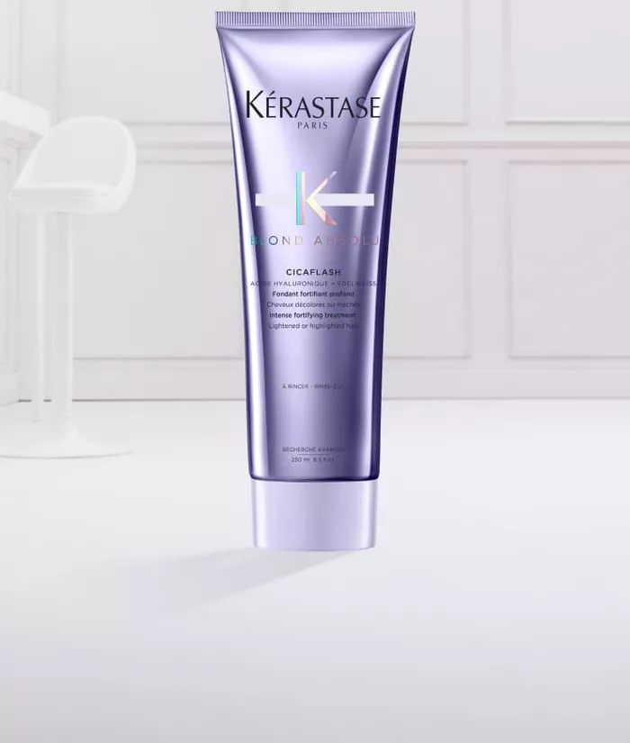 Ein lila Tubus von Kerastase „Blond Absolu“ Cicaflash mit einer cremigen, blauen Haarpflegeformulierung, die aus der geoffneten Tube herausquillt.