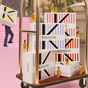 A cart stacked with colorful Kerastase product boxes and small bottles, with a person in the background carrying a large box. The setting features a pink backdrop.