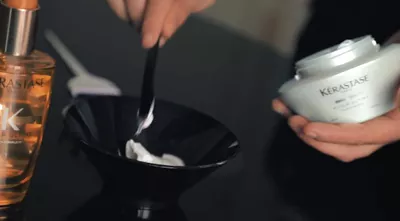 A hand holds a black mixing bowl containing a white cream, with a black spatula for mixing; a bottle of hair oil is visible in the background.