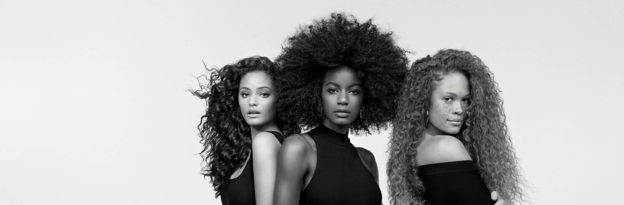A black-and-white portrait of three women standing side by side, each showcasing different hairstyles: one with loose curls, one with a large afro, and one with wavy red hair. They are wearing sleeveless black tops and are posed against a plain background.
