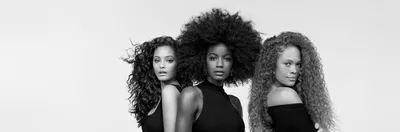 A black-and-white portrait of three women standing side by side, each showcasing different hairstyles: one with loose curls, one with a large afro, and one with wavy red hair. They are wearing sleeveless black tops and are posed against a plain background.