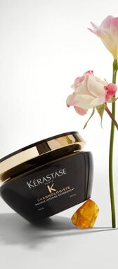 A jar of Kerastase Chronologiste hair mask with an elegant black design and gold accents, positioned beside a delicate pink rose and a yellow crystal on a soft, neutral background.