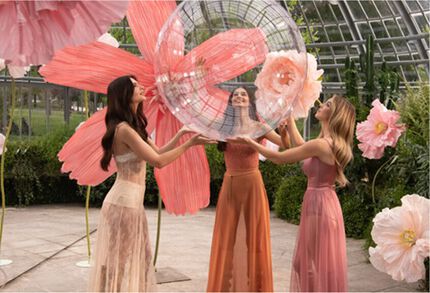 Drei Frauen in fließenden Kleidern interagieren mit transparenten Blasen und riesigen rosa Blumen in einer uppigen Umgebung.
