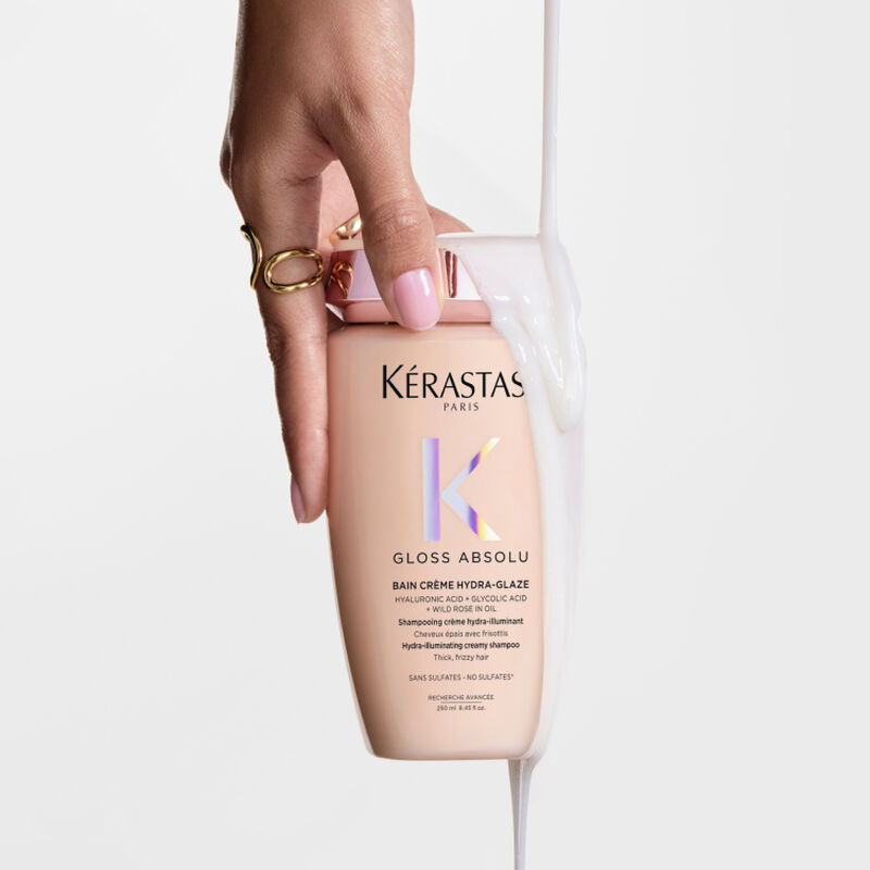 Eine Hand halt eine Flasche Kerastase Gloss Absolu Shampoo, uber die eine cremige weiße Flussigkeit gegossen wird.