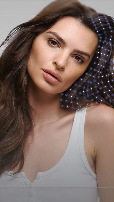 Eine Frau mit langen, dunklen Haaren, die ein weißes Oberteil tragt, schaut direkt in die Kamera. Ihre Gesichtszuge sind sanft und sie hat einen neutralen Ausdruck. Hinter ihr ist ein verschwommener Hintergrund.