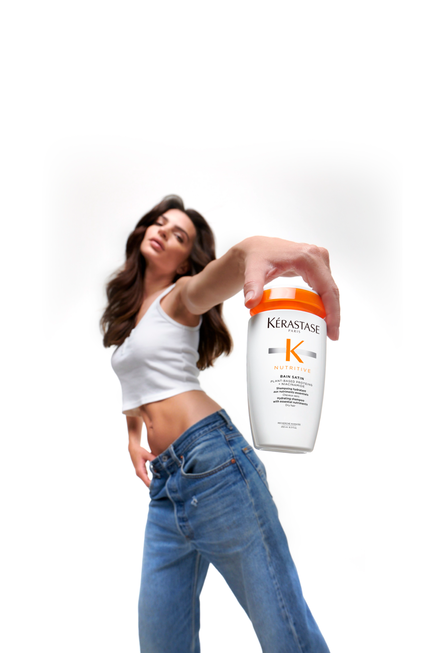 A model in a white tank top is holding a jar of Kerastase Nutritive hair care product, posing confidently against a white background.