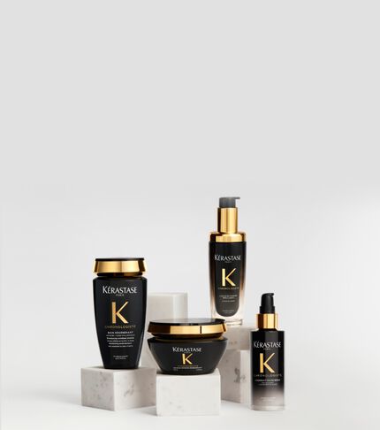 Haarpflegeprodukte der Serie Kerastase Chronologiste in schwarz-goldenen Flakons, elegant arrangiert auf weißen Marmorsockeln vor hellem Hintergrund.