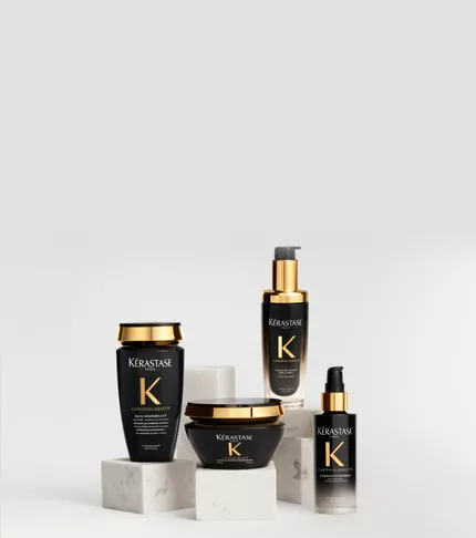 Haarpflegeprodukte der Serie Kerastase Chronologiste in schwarz-goldenen Flakons, elegant arrangiert auf weißen Marmorsockeln vor hellem Hintergrund.