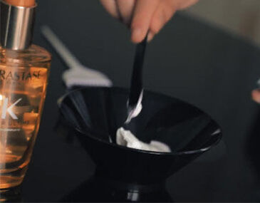 A hand holds a black mixing bowl containing a white cream, with a black spatula for mixing; a bottle of hair oil is visible in the background.