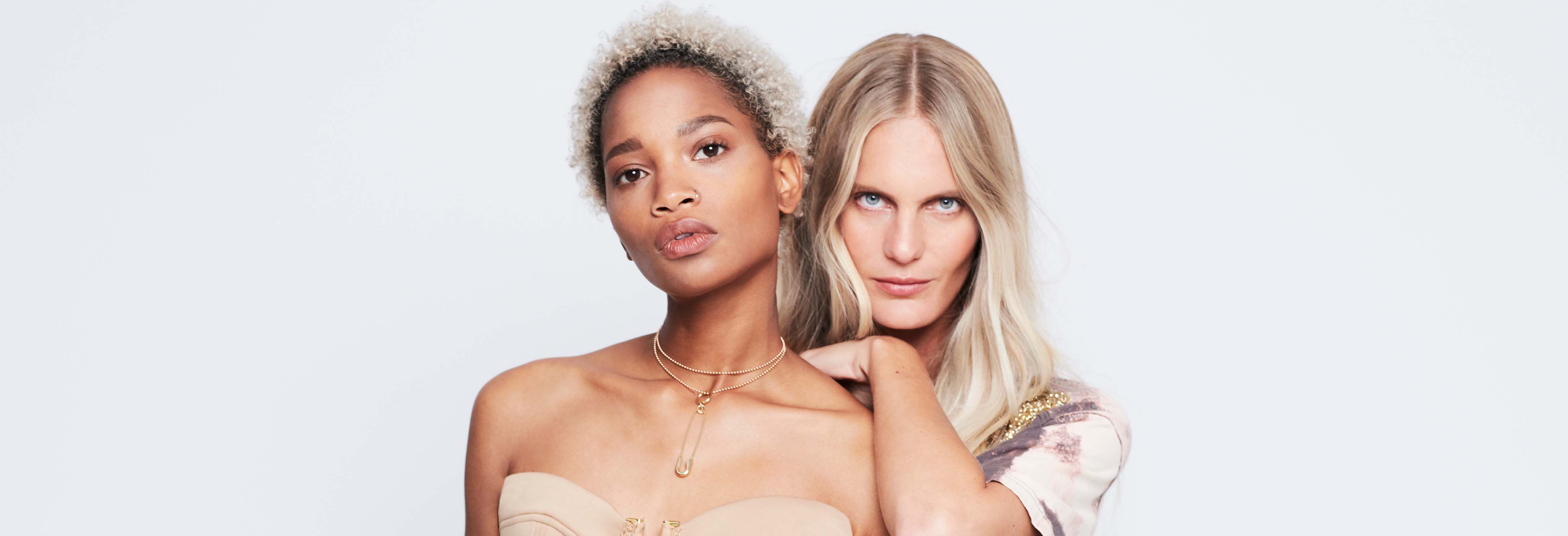 Two women pose closely together against a plain white background. One has curly blonde hair and is wearing a strapless top, while the other has long straight hair and a patterned shirt. They both have confident expressions.