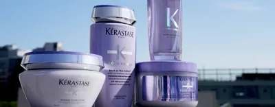 Four Kerastase hair care products in purple containers are displayed against a bright background, featuring labels with the brand name and product details.