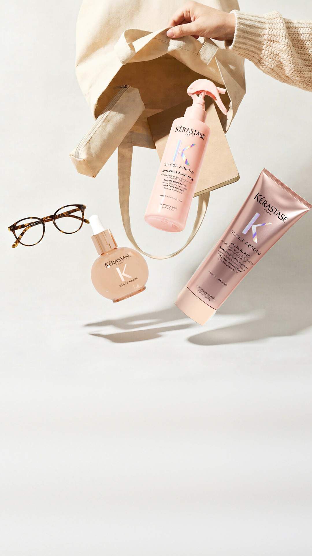 Drei Flaschen von Kerastase Haarpflegeprodukten (Elixir Ultime, Genesis und Nutritive) sind auf einer weißen Oberflache neben einer Vintage-Kamera und mehreren weißen Pfingstrosen mit grunen Blattern angeordnet. Die goldene Elixir Ultime Flasche mit schwarzem Pumpverschluss ist links, die pinke Genesis Flasche mit silberner Pipette ist in der Mitte, und die rot-orange verlaufende Nutritive Flasche mit silberner Pipette ist rechts. Die silberne Kamera mit ihrem schwarzen Band befindet sich links von den Produkten.