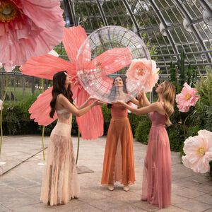 Three women in elegantly designed dresses hold a large, transparent sphere amid oversized pink flowers in a greenhouse setting.