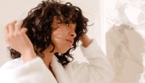 A person with curly hair adjusts their hair while wearing a white bathrobe, standing in a softly lit bathroom.