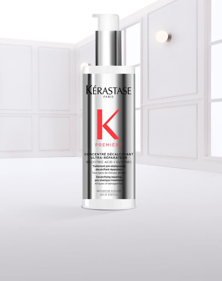 Ein transparentes Flaschchen mit einem weißen Pumpspender und silberner Kappe, das Kerastase auf dem Etikett tragt. Es sieht aus, als ob es mit Wasser benetzt ist, und Tropfen laufen von der Flasche.
