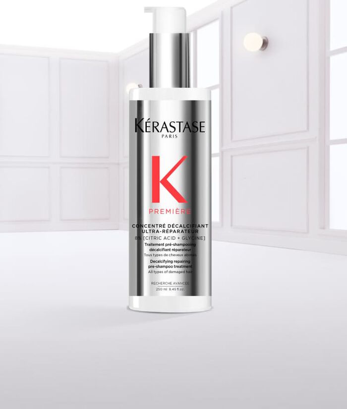 Ein transparentes Flaschchen mit einem weißen Pumpspender und silberner Kappe, das Kerastase auf dem Etikett tragt. Es sieht aus, als ob es mit Wasser benetzt ist, und Tropfen laufen von der Flasche.