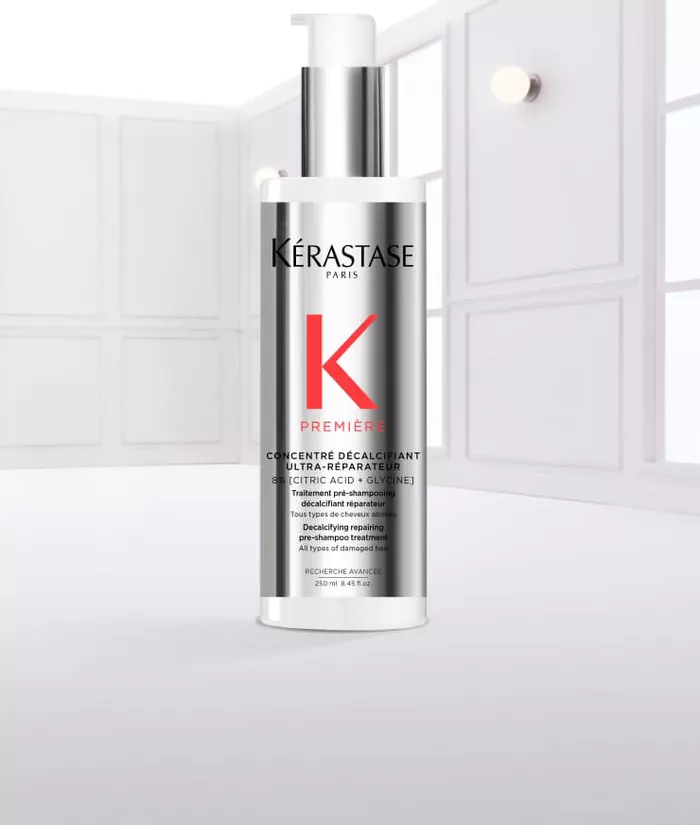 Ein transparentes Flaschchen mit einem weißen Pumpspender und silberner Kappe, das Kerastase auf dem Etikett tragt. Es sieht aus, als ob es mit Wasser benetzt ist, und Tropfen laufen von der Flasche.