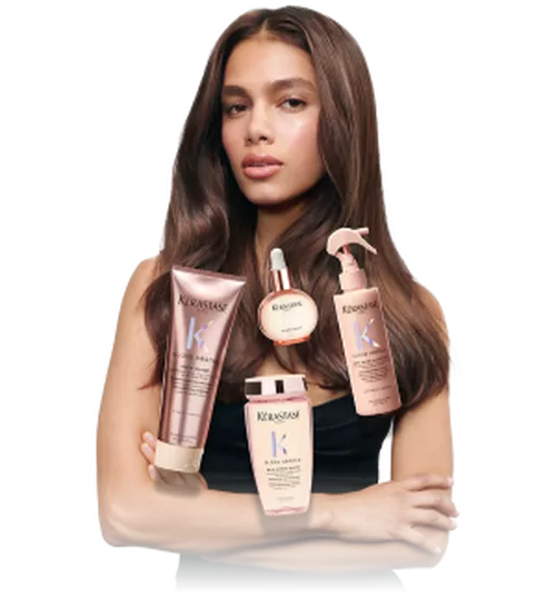 A model with long, smooth hair stands confidently, holding several Kerastase hair care products.