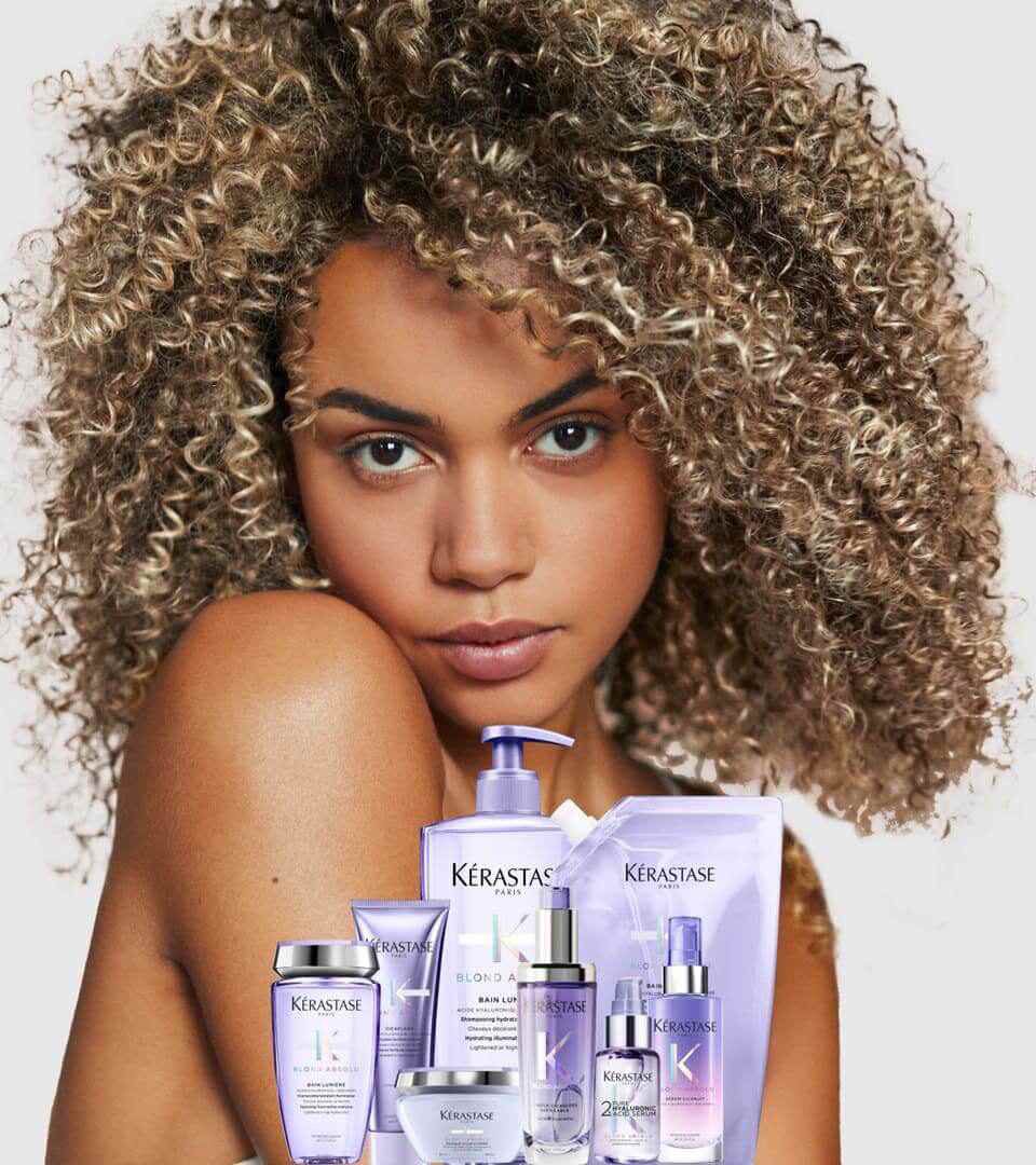 A woman with curly hair poses with her arms crossed, surrounded by various Kerastase hair care products in purple packaging.