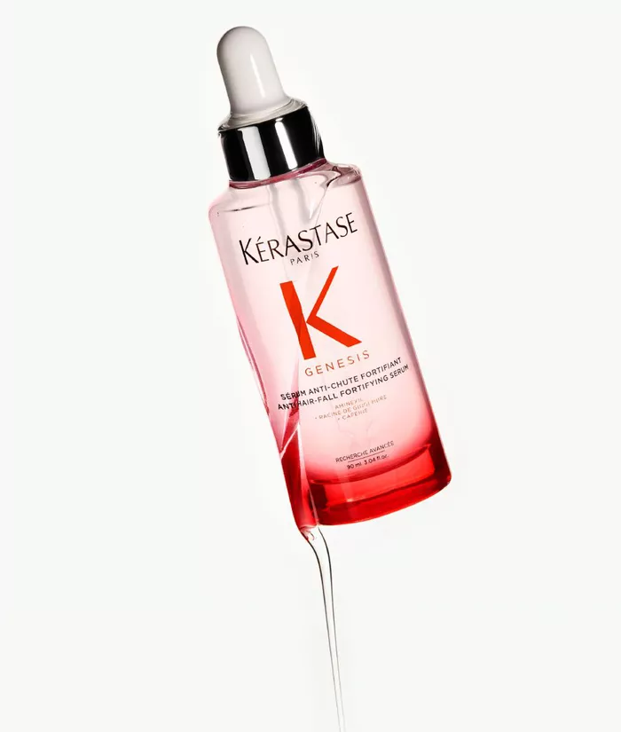 Eine Flasche Kerastase Genesis Haarserum mit Pipette, auf einem hellen Hintergrund, umgeben von einigen Flussigkeitstropfen.