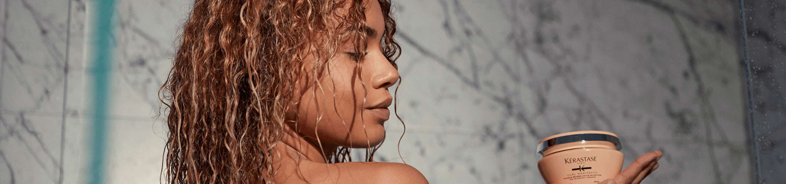 A woman with curly hair holds a jar of hair treatment while standing in a shower, surrounded by marble tiles.