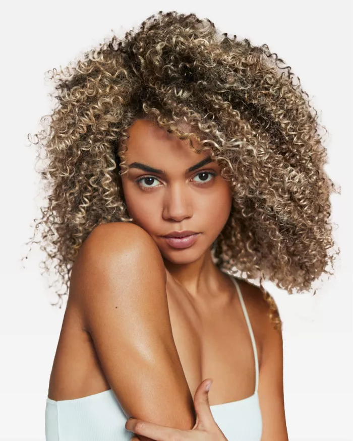 Eine junge Frau mit lockigem, blondem Haar schaut direkt in die Kamera und umarmt leicht ihren Arm. Sie hat einen naturlichen, frischen Look.