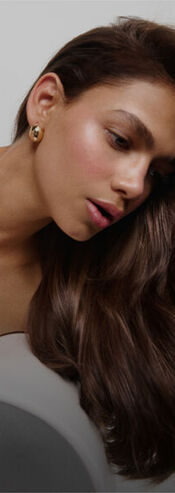 A side-profile portrait of a woman with long, wavy brown hair and a soft complexion, wearing a subtle golden earring. She appears contemplative, slightly leaning forward.