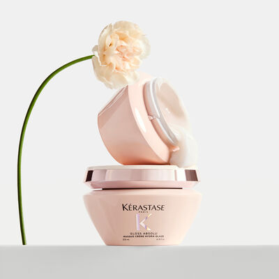 Zwei Dosen Kerastase Gloss Absolu Haarmaske mit herausfließender cremeweißer Textur und einer blassrosa Nelke.