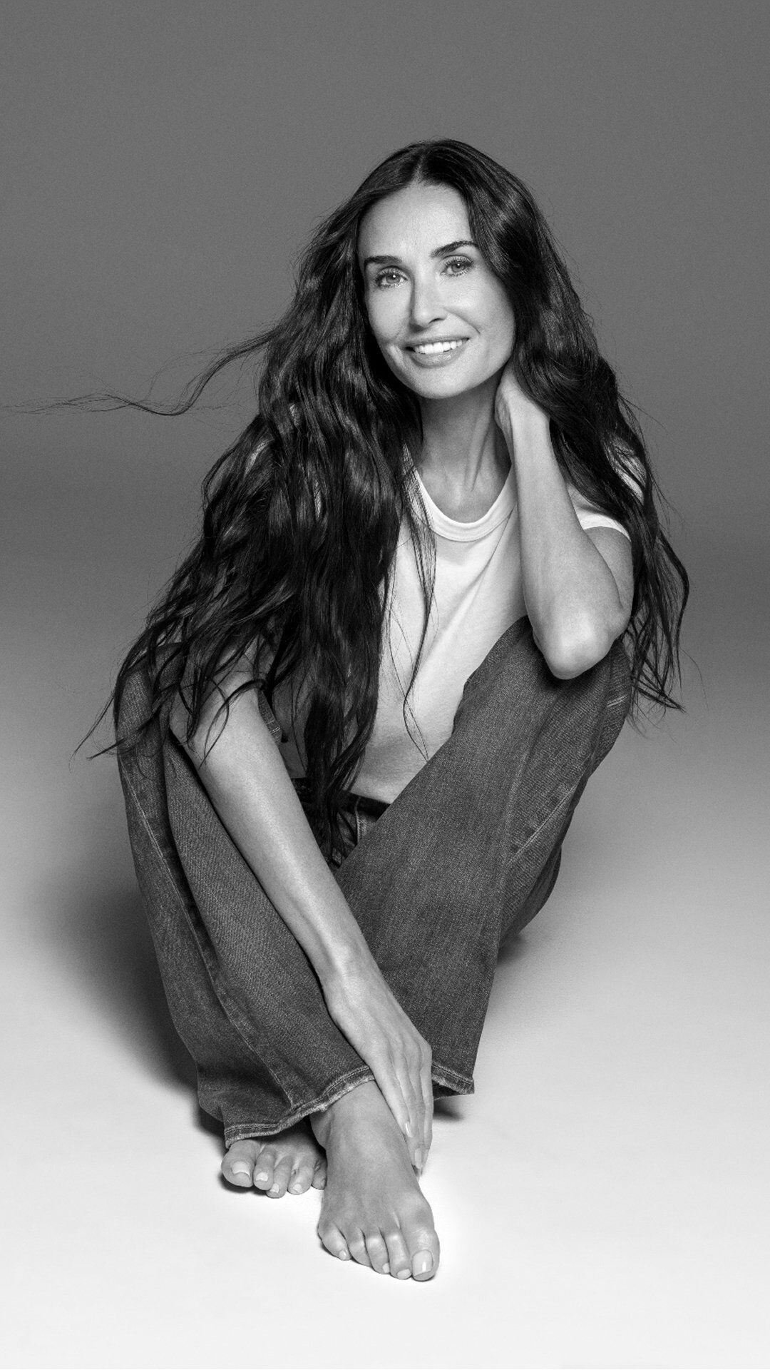 Demi Moore im Schneidersitz sitzend mit einem breiten, strahlenden Lacheln.​