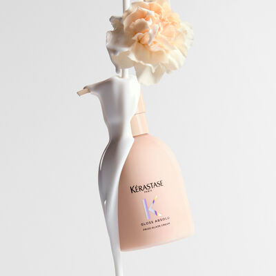 Eine Flasche Kerastase Gloss Absolu Frizz-Glaze Creme, uber die eine cremige weiße Flussigkeit gegossen wird, mit einer hellen Nelke an der Spitze.