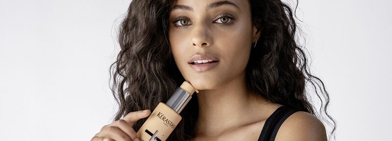 Eine Frau mit lockigem Haar halt eine Foundation-Flasche in der Hand und schaut direkt in die Kamera. Sie tragt ein einfaches schwarzes Top und hat einen naturlichen Look.