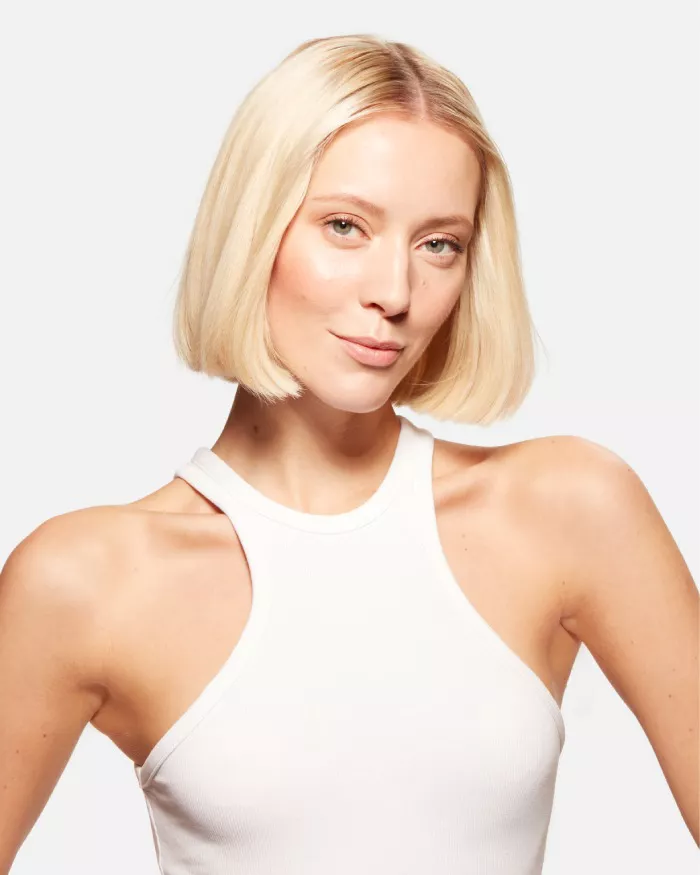 Eine blonde Frau mit schulterlangen Haaren tragt ein armelloses, weißes Oberteil und schaut direkt in die Kamera.