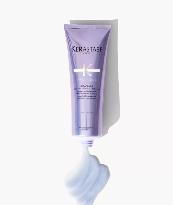 Ein lila Tubus von Kerastase „Blond Absolu“ Cicaflash mit einer cremigen, blauen Haarpflegeformulierung, die aus der geoffneten Tube herausquillt.