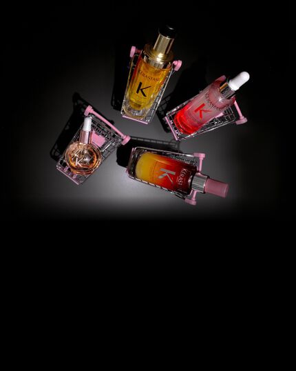 Vier Kerastase Haarpflegeprodukte in rosa Mini-Einkaufswagen auf schwarzem Hintergrund: das Nutritive Serum Nuit 8H, das Elixir Ultime L'Huile Originale, das Serum Anti-chute Fortifiant de Genesis und die Glaze Drops.