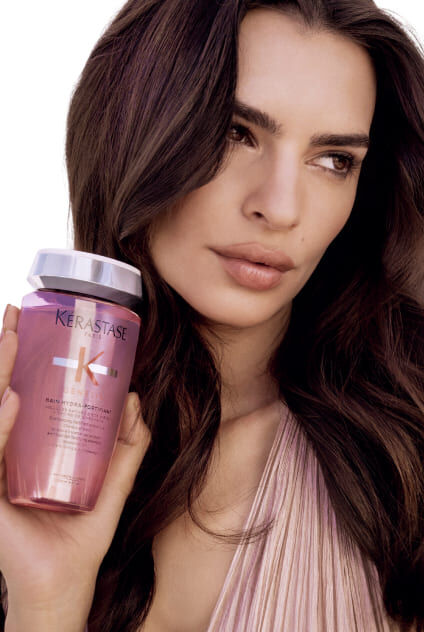 Frau halt eine transparentrosa Flasche Kerastase Genesis Bain Hydra-Fortifiant, das Produkt prasentierend.