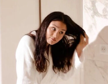 A woman in a white bathrobe is brushing her long hair while looking to the side. The background features a light-colored wall and a small electrical outlet.