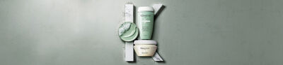 Three jars of skincare products in soft green and white colors are displayed against a textured gray background, with one jar partially opened and a small amount of product shown.