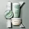 Three jars of skincare products in soft green and white colors are displayed against a textured gray background, with one jar partially opened and a small amount of product shown.