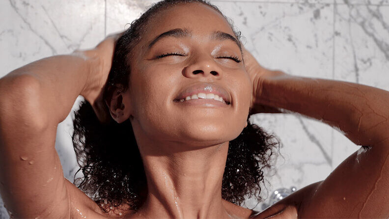 A young woman with curly hair smiles gently while washing her hair in the shower, water glistening on her skin against a marble background.