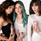 Three women pose together, holding various hair care products, with a neutral background. Each woman has a distinct hairstyle: one with curly hair, another with straight hair and teal streaks, and the third with sleek, straight hair.