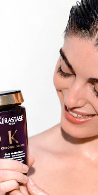A person with wet hair smiles while holding a jar of Kerastase hair care product.