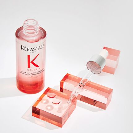 Eine Flasche Kerastase Genesis Serum mit Pipette, umgeben von geometrischen, rosafarbenen Blocken.