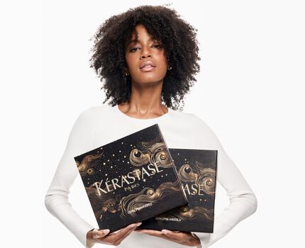 A young woman with curly hair holds two boxes with intricate designs against a white background. She is wearing a white long-sleeve shirt.