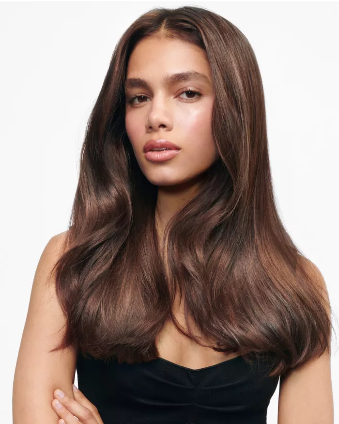 Ein junges Model mit langen, glanzenden Haaren und einem naturlichen, strahlenden Makeup, das in die Kamera schaut. Sie tragt ein schwarzes Oberteil und hat eine entspannte Haltung.