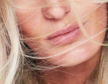 Close-up of a woman's face with blonde hair blowing across her lips, highlighting soft skin and natural beauty.