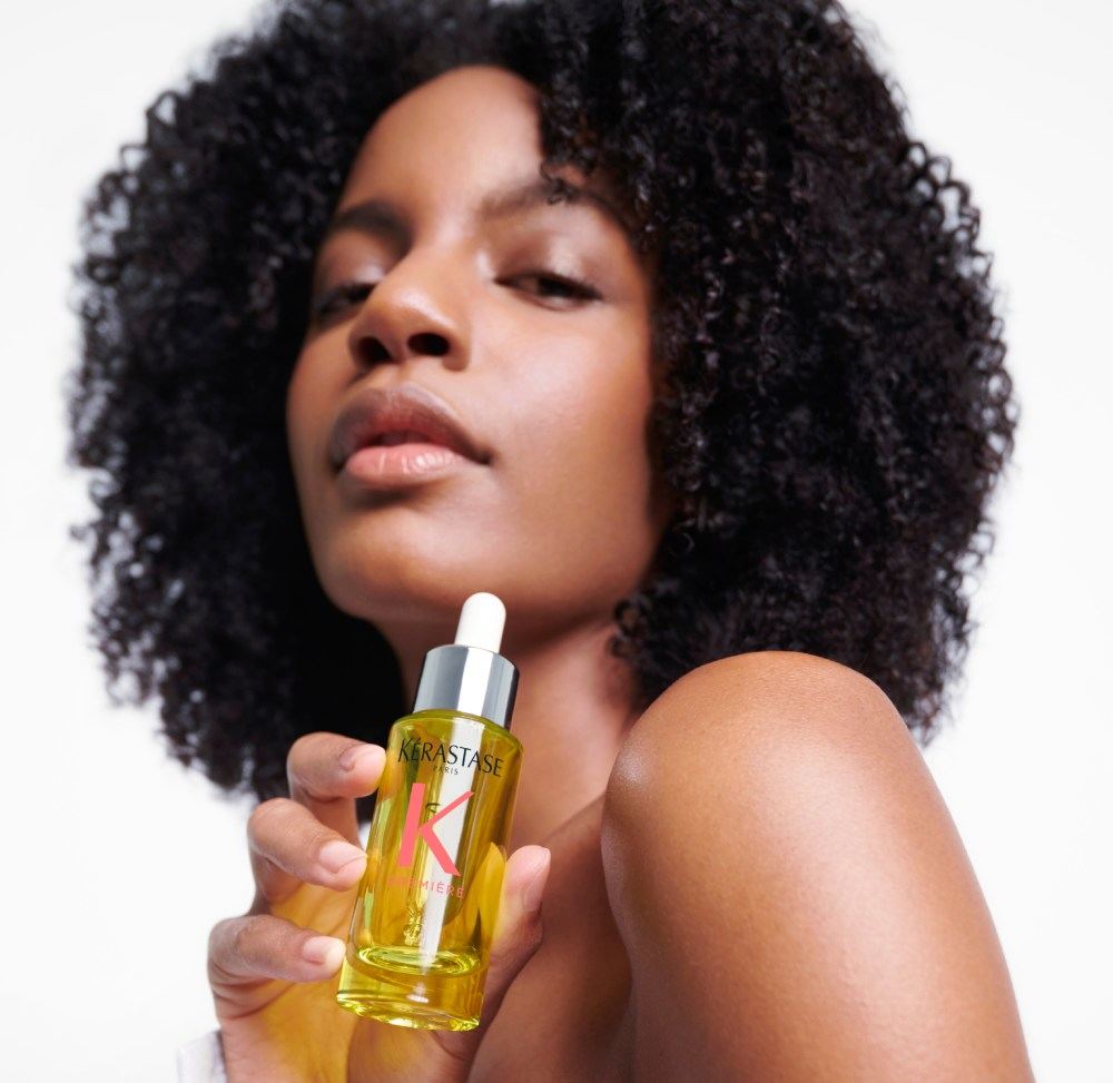 Ebonee Davis mit ihrem wunderschönen lockigen Haar hält das beste Haaröl für geschädigtes Haar Première Huile Gloss Réparatrice in ihren Händen.