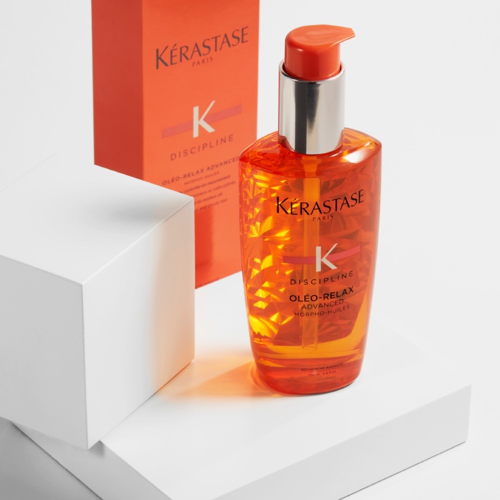 Das Haaröl Discipline Oleo Relax wird auf einer einfachen weißen Plattform auf einem rein grauen Hintergrund präsentiert, aus dem sich seine leuchtend orange Farbe glitzernd hervorhebt.