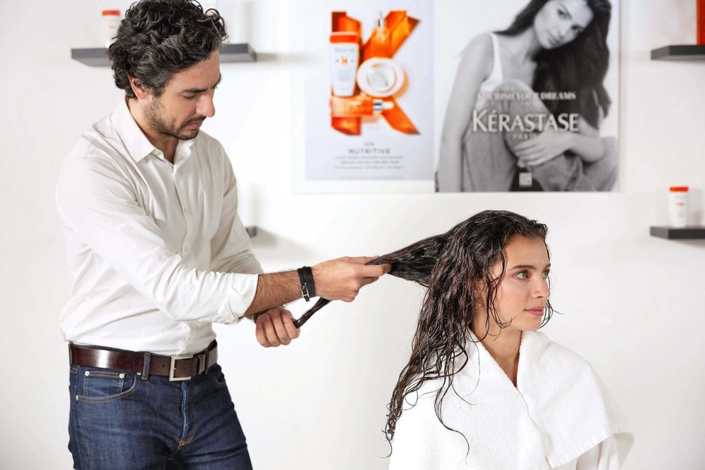 Ein Friseur stylt das nasse Haar einer Frau in einem modernen Salon, im Hintergrund Produkte von Kérastase.