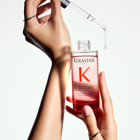 Nahaufnahme von Händen, die eine Flasche Kérastase Genesis Stärkendes Serum halten, mit einer Pipette, aus der ein Tropfen des Produkts fallen wird.